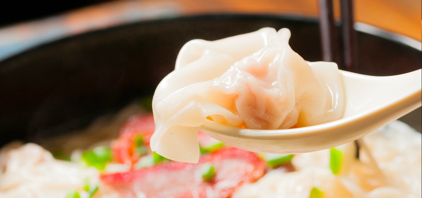 A wonton on a Chinese soup spoon, with chopsticks and a hearty bowl of soup in the background.